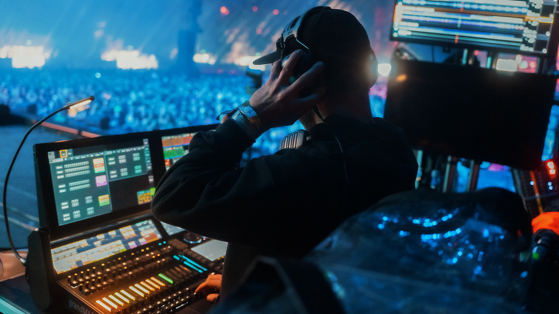 A DJ wearing headphones and a baseball cap operating sound and lighting equipment at a concert, with a large crowd and stage lights in the background.