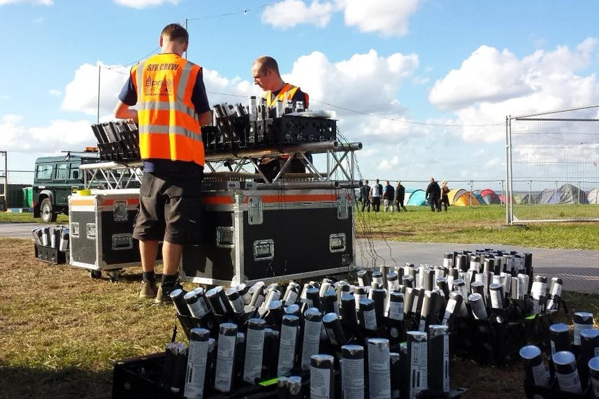 Two men wearing safety vests working at a DJ booth with audio equipment outdoors, with many cans placed on the ground and in the background, tents and a fence with clouds in the sky.