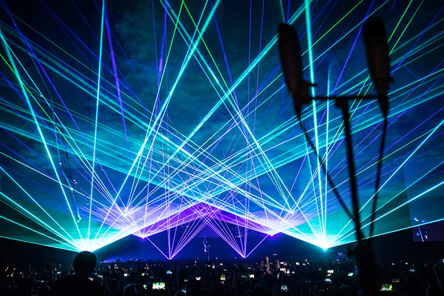 A concert or show with numerous blue and green laser lights crossing in the air, with a crowd of people in the foreground taking photos and videos.