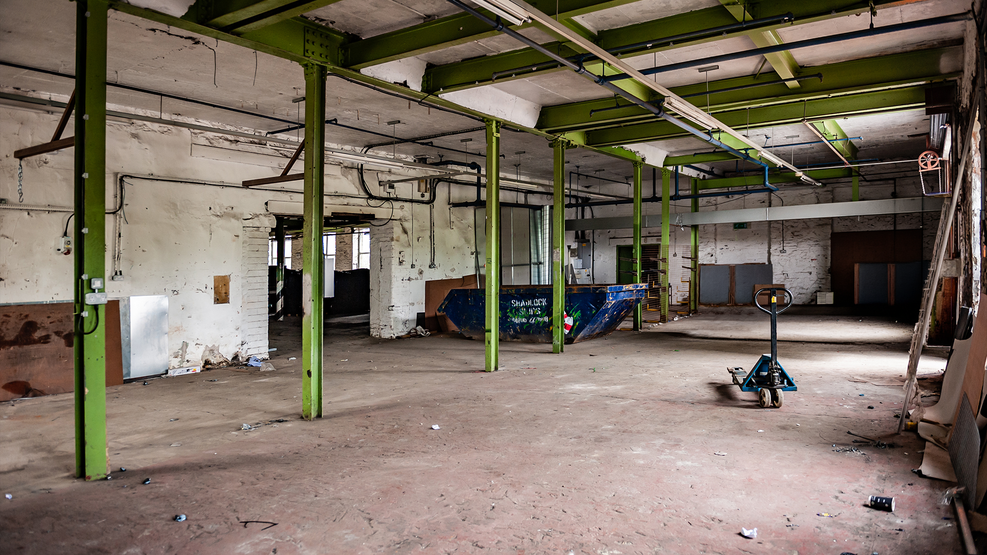 An empty, unfinished industrial or warehouse space with exposed green steel beams, white brick walls, and a concrete floor. There is a small blue cart or pallet jack in the foreground, and some construction or renovation debris scattered around.