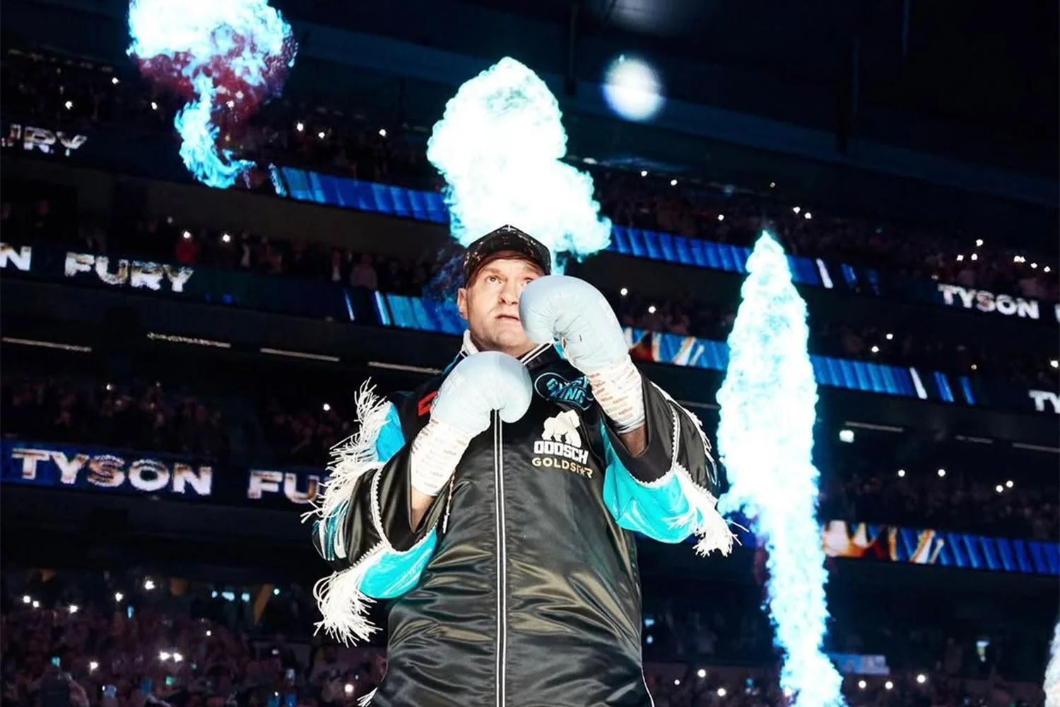 A professional boxer standing in a boxing ring with pyrotechnics firing behind him during a match, wearing boxing gloves and a jacket with a logo.