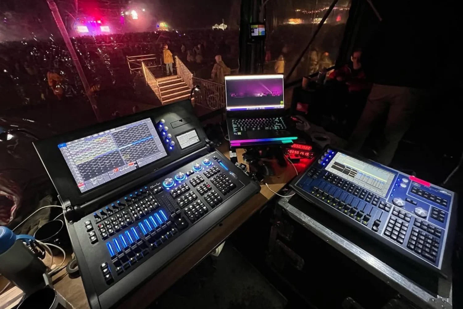 DJ booth with multiple digital mixing consoles, laptops, and controllers at a nighttime event with a crowd outside on a lit stage.