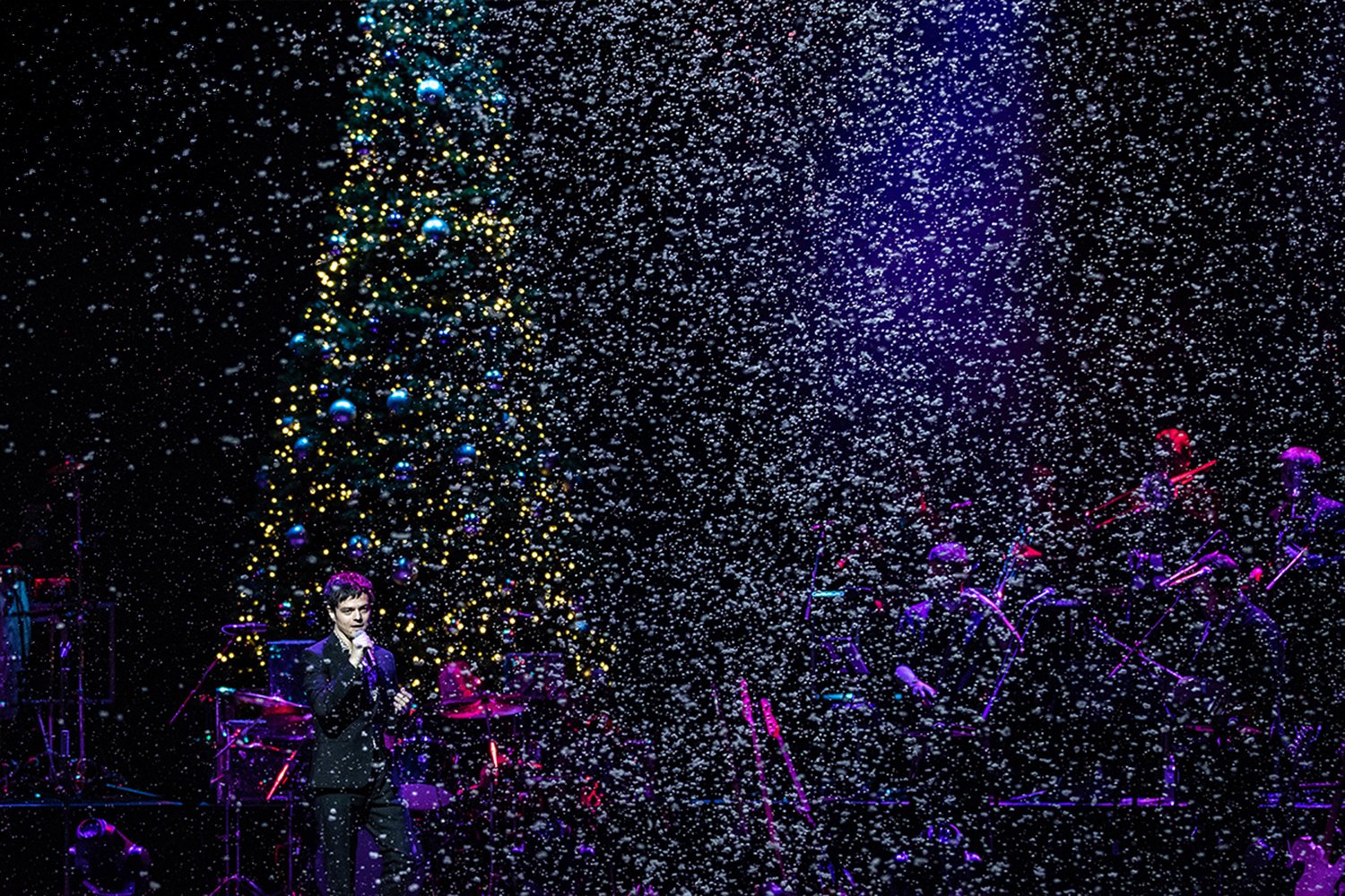 A performer singing on stage with a Christmas tree decorated with lights and ornaments in the background, surrounded by falling snow or confetti.