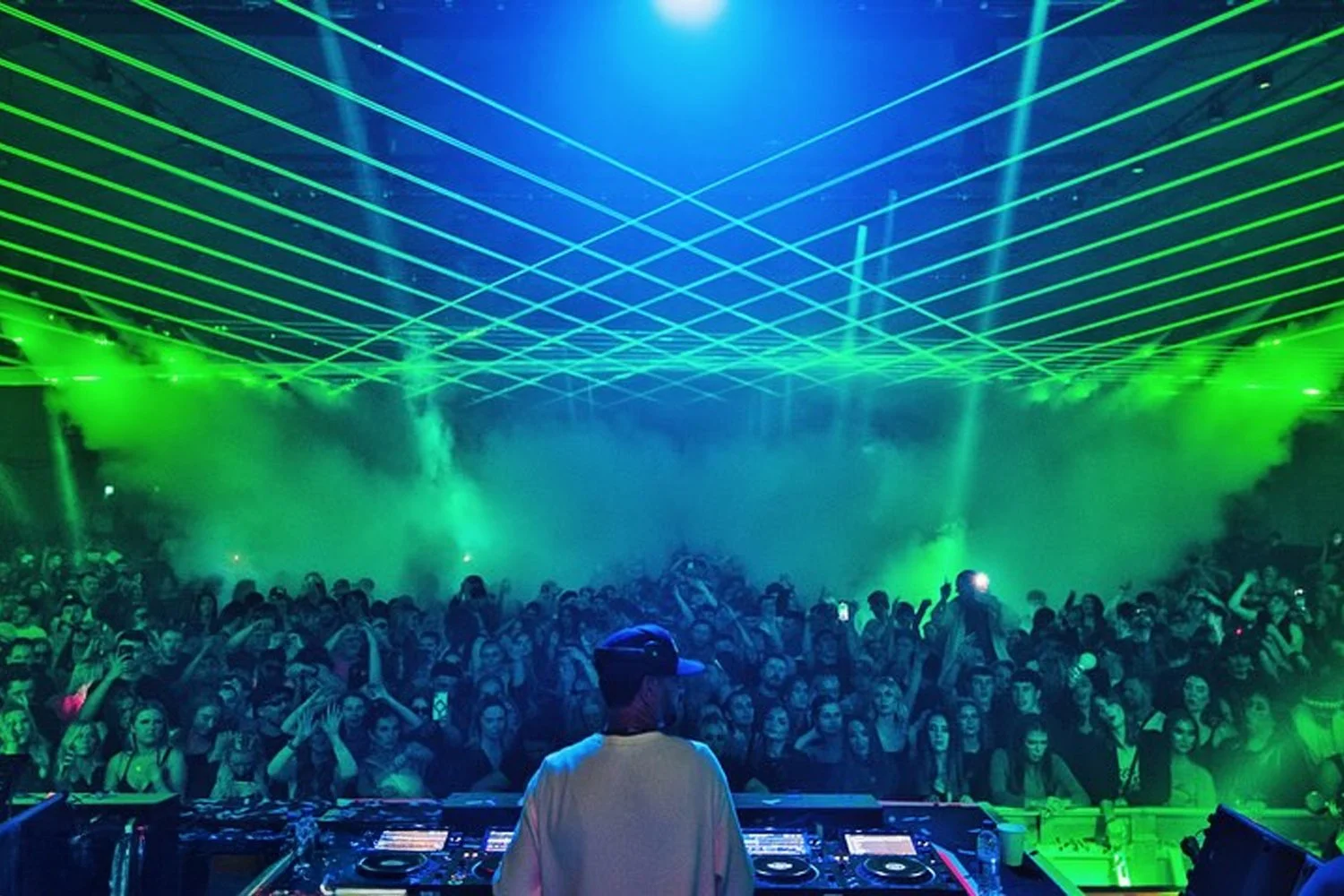A DJ performing at a nightclub with a crowd dancing. Green laser lights and smoke effects create a vibrant atmosphere.