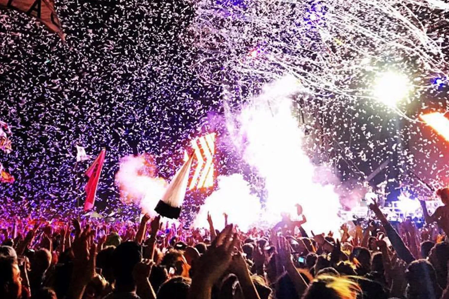 Crowd of people celebrating with confetti, fireworks, and bright lights at a nighttime event.