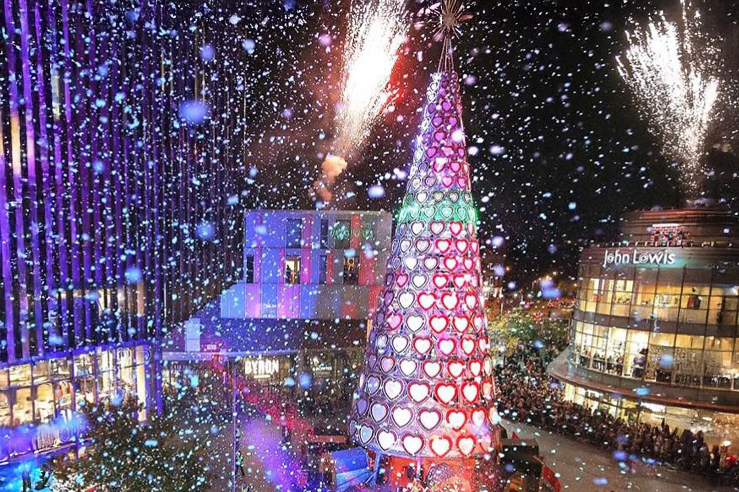 Nighttime scene with colorful Christmas tree decorated with illuminated heart-shaped lights, surrounded by people. Fireworks light up the sky above a shopping area with stores such as John Lewis and BYTON, decorated with holiday lights and decorations.