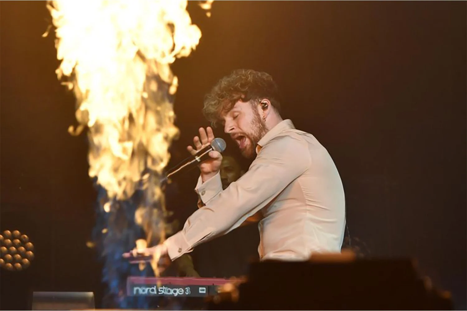 A man with curly hair and beard, wearing a light-colored shirt, performing passionately at a music concert, singing into a microphone with flames on a fire prop nearby, and a keyboard labeled 'nord stage 3' in front of him.