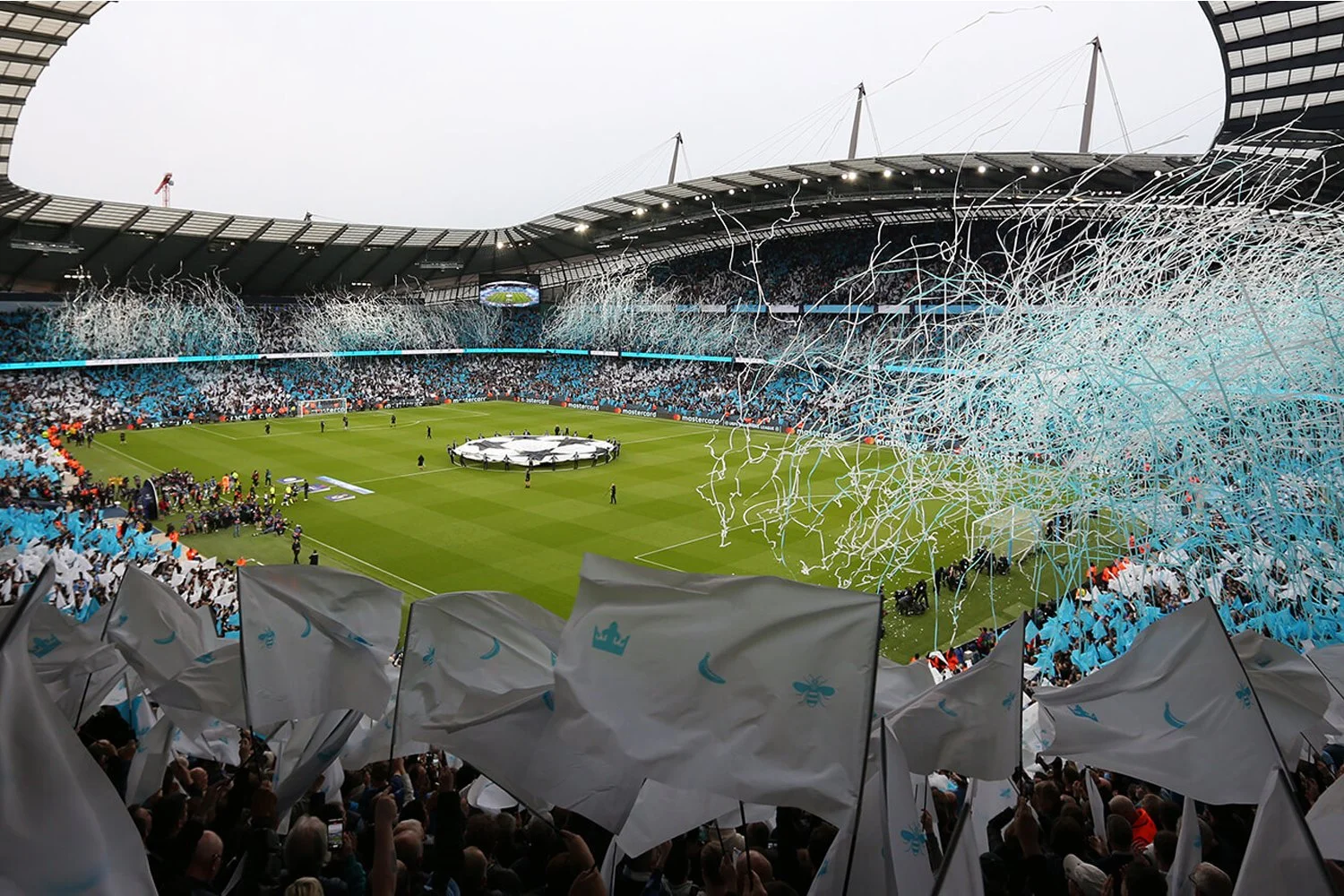 A packed soccer stadium with fans holding white flags and blue decorations, confetti falling, and preparations for a UEFA Champions League match.
