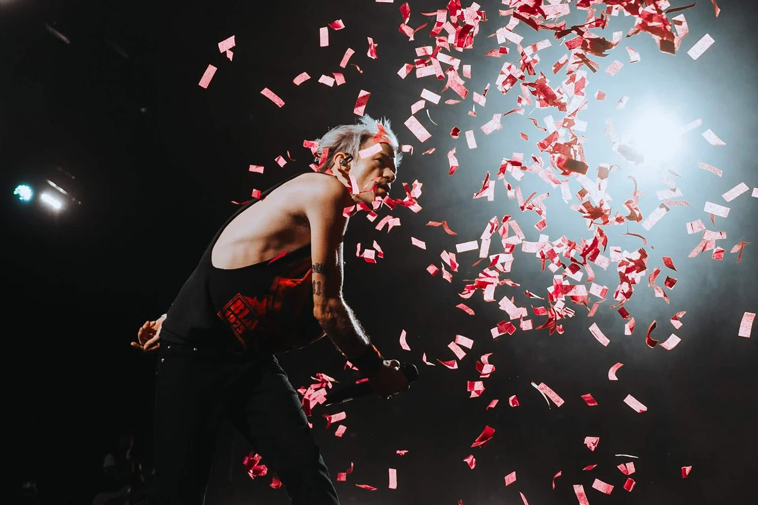 A man on stage during a concert, surrounded by falling red and white confetti, holding a microphone, with bright stage lighting.