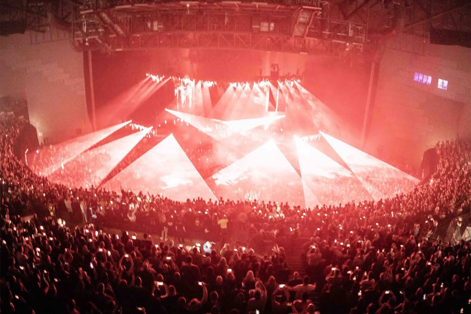 Crowd attending a concert or music event in a large indoor arena, with red stage lighting and visual effects.
