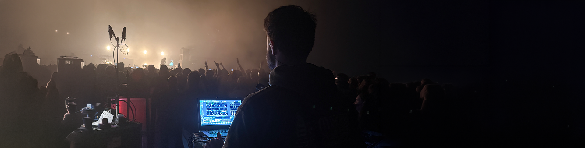 A DJ is performing at a concert or music event at night, with a crowd of people in front of him, and the scene is lit with stage lights and fog.