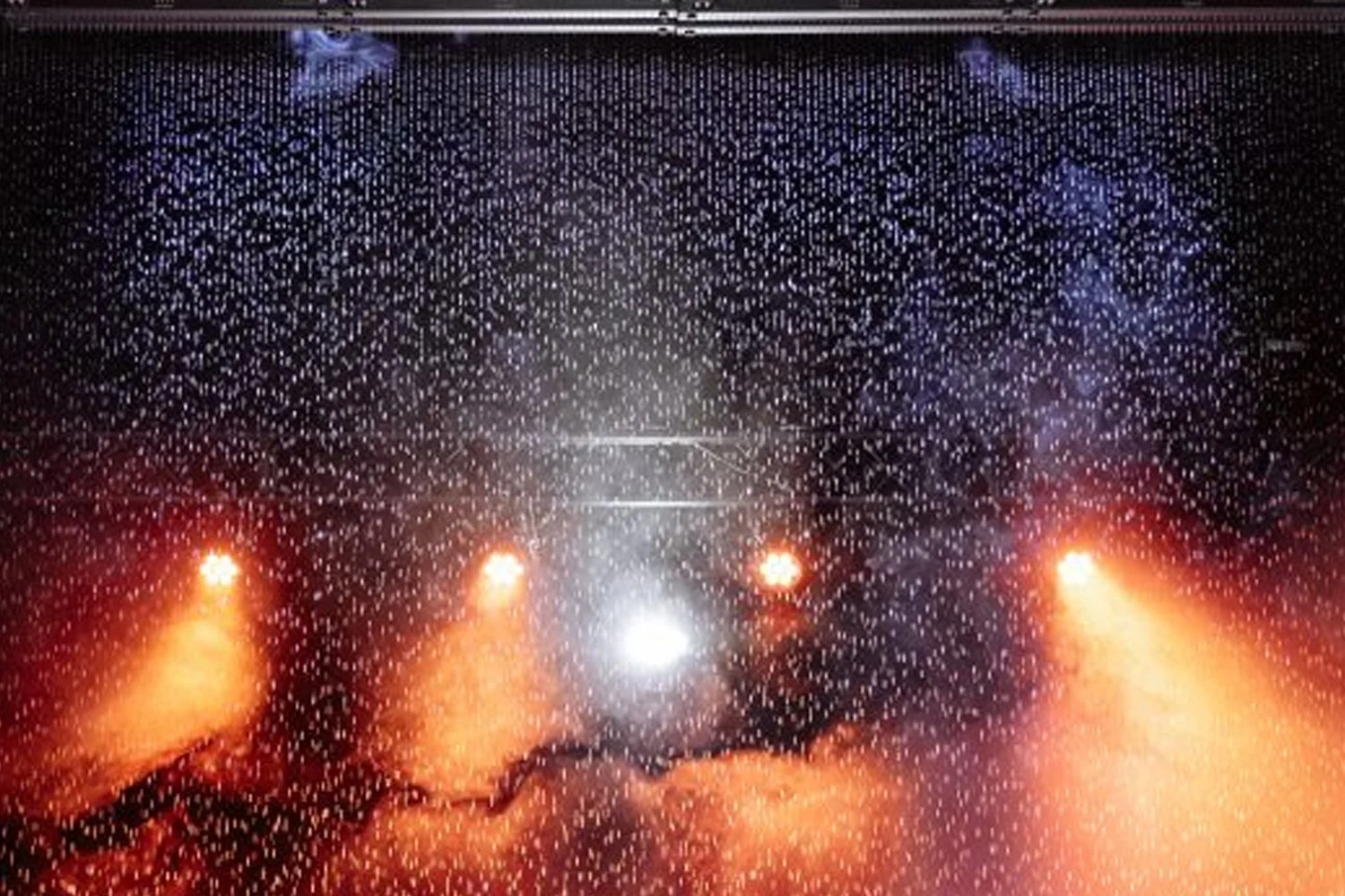 A view through a rain-covered window with three orange stage lights, colorful smoke or fire effects, and a bright white light in the background.