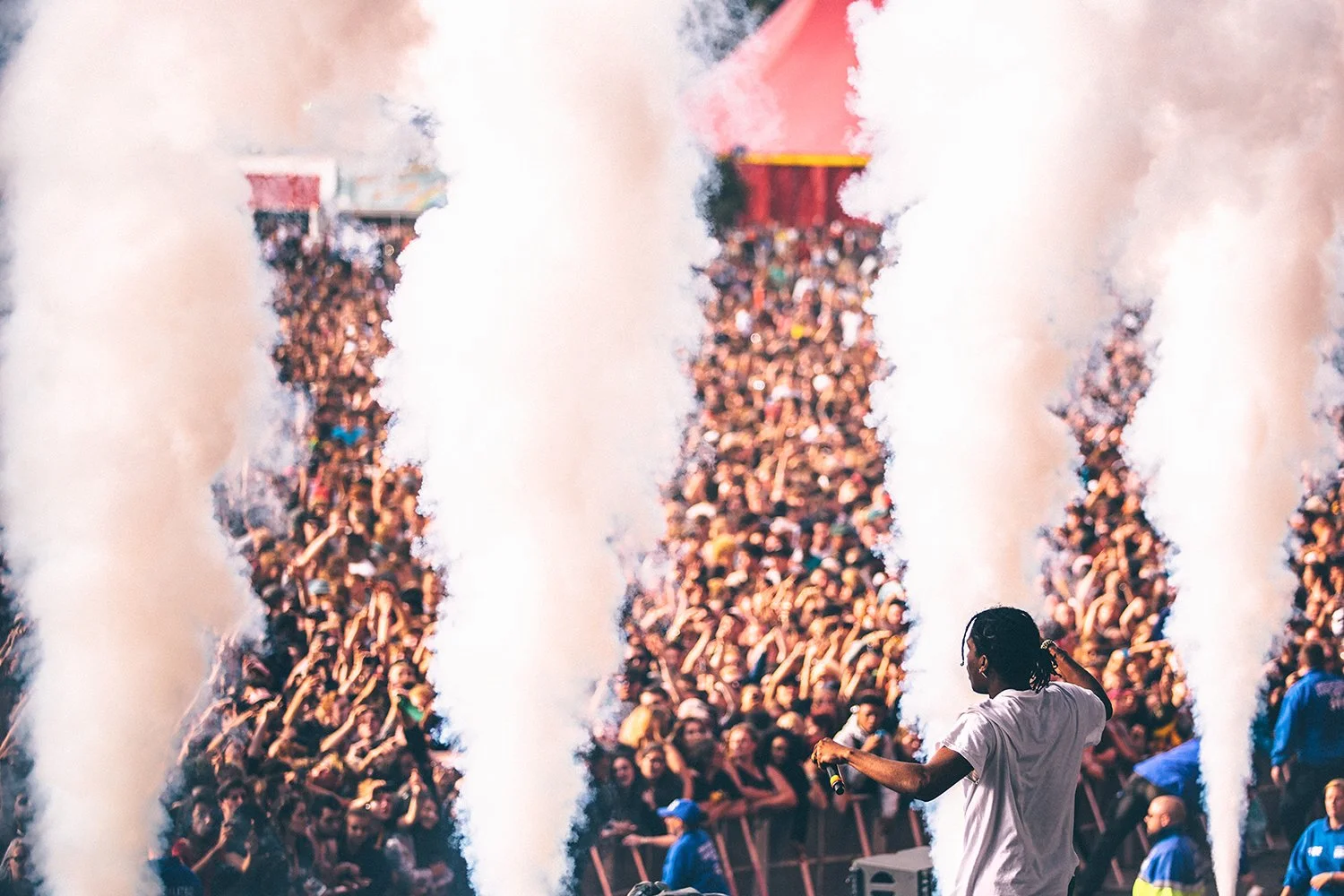 A large crowd of people at a concert or festival, with smoke or fog effects and a performer on stage in the foreground.