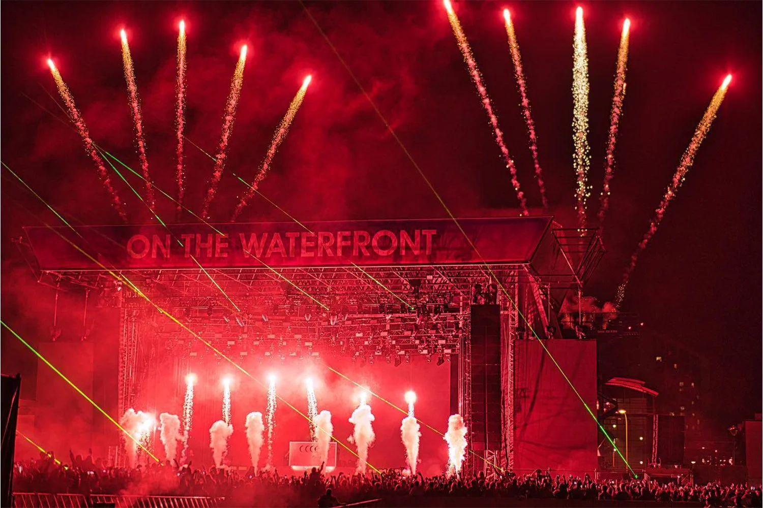 Nighttime concert on a waterfront with fireworks, smoke, and laser lights illuminating the stage and crowd, with a sign that says 'ON THE WATERFRONT'.