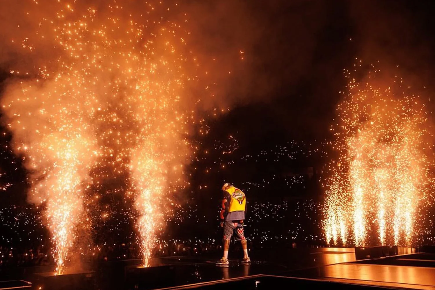 A performer on stage surrounded by fireworks and sparks during a nighttime outdoor show.