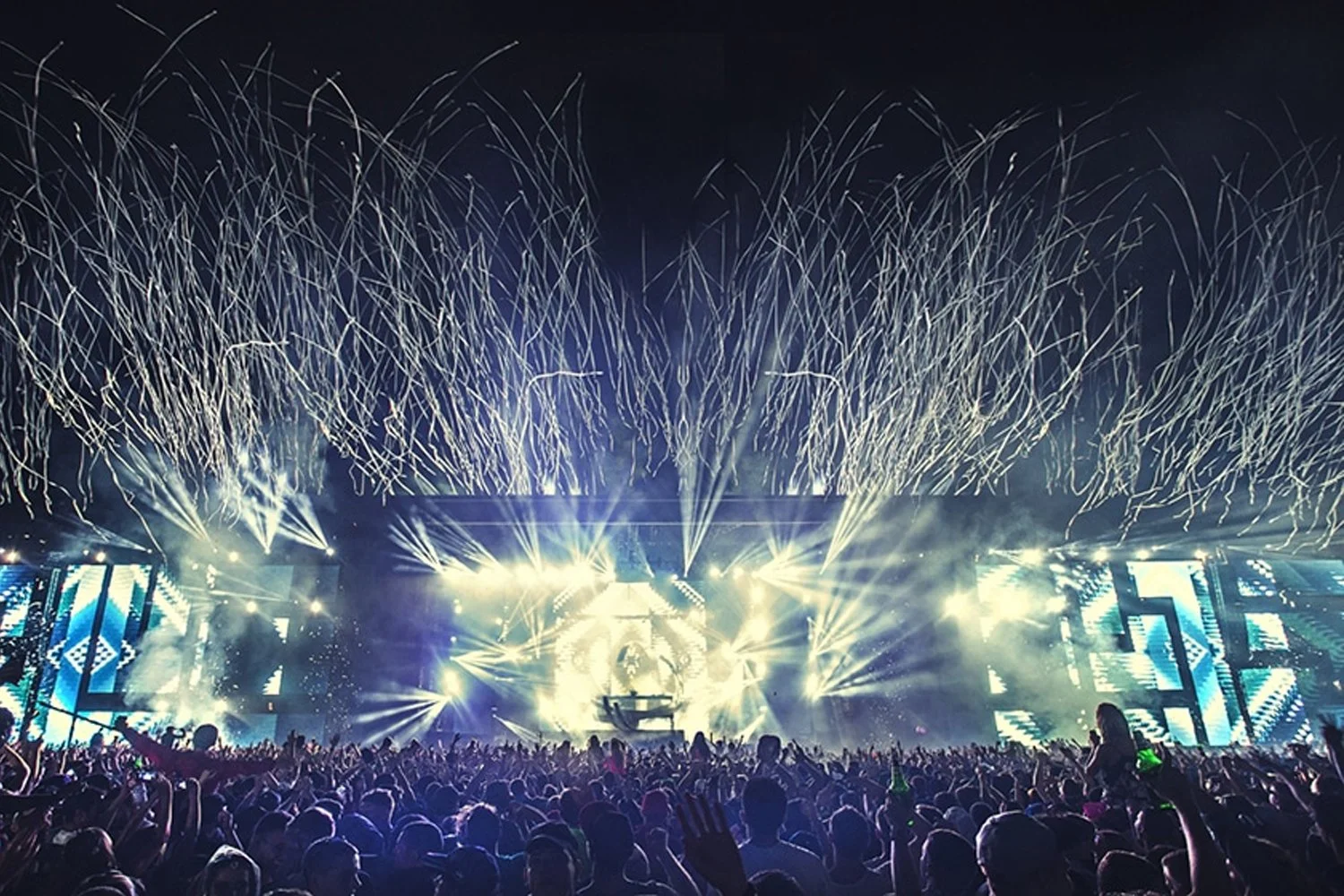 Crowd at a large electronic dance music concert with colorful lights, smoke, and fireworks in the night sky.