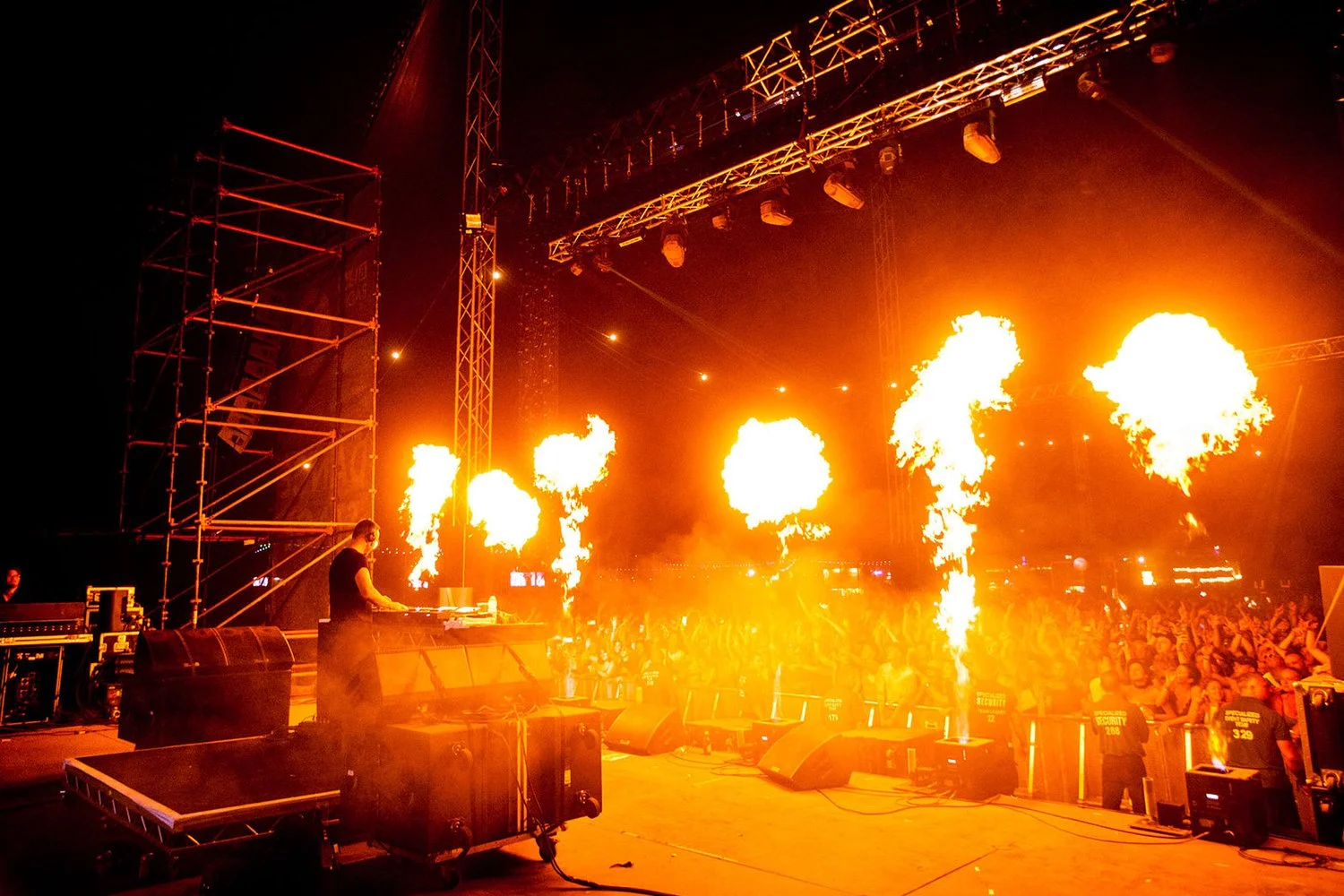 A DJ performs on stage at a music concert with fire pyrotechnics shooting upwards behind him, and a crowd of people dancing in front.