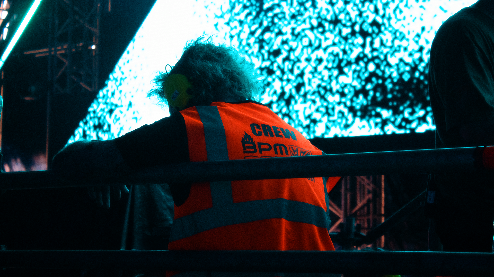A person with curly hair wearing an orange and gray safety vest labeled 'CREW' and noise-canceling headphones, leaning on a barrier at a concert or event with a large LED screen displaying blue and white patterns in the background.