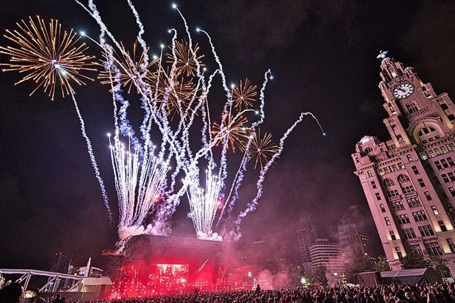 Fireworks display over a large crowd at night with a historic building on the right and a stage with bright lights and a LED screen at a waterfront event.