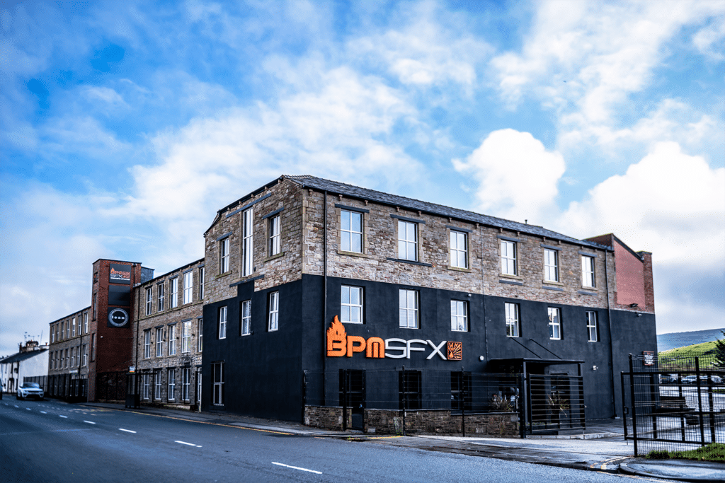 A three-story brick building with a black-painted lower section, featuring the 'BPM SFX' sign on the front, situated on a street with a clear sky and some clouds overhead.