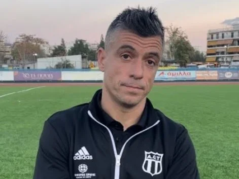 A man with short dark hair and a slight smirk, wearing a black adidas sports jacket with a logo reading A.C.T. and standing on a grassy sports field with a track, trees, and buildings in the background.