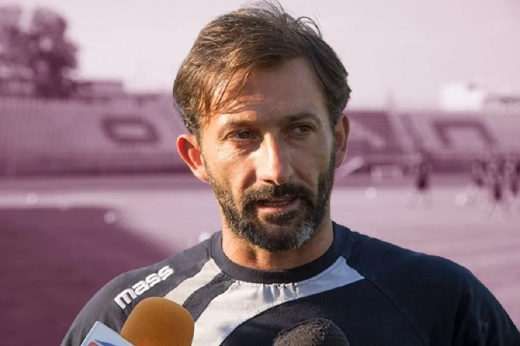 A man with brown hair and a beard speaking into a microphone, outdoors in front of an aircraft.