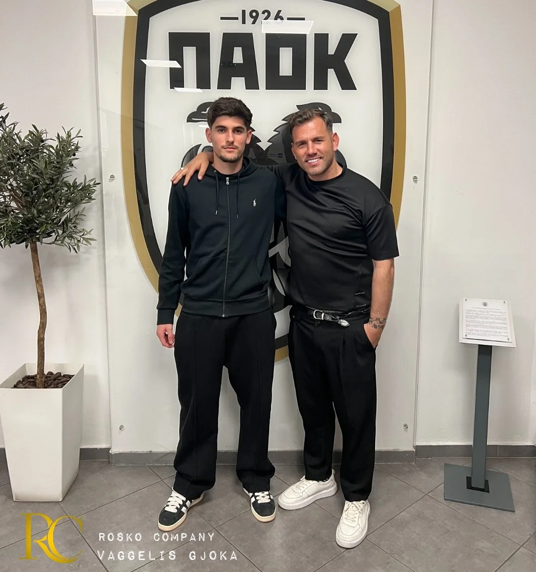 Two men standing together, one with his arm around the other's shoulder, in front of a large shield-shaped logo that says '1926' and other words. The man on the left has dark hair and is wearing a black zip-up hoodie and black sweatpants, while the man on the right has short hair, a beard, and is dressed in a black t-shirt and black pants. A potted plant is on the left side of the image, and a small sign on a stand is on the right side.