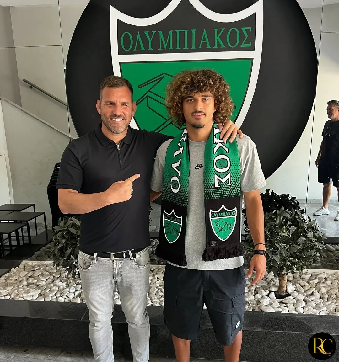 Two men standing in front of a large Olympiakos logo, one pointing at the other. The man on the right wears a scarf with the Olympiakos emblem, and the man on the left is smiling, wearing a black polo shirt and light jeans.