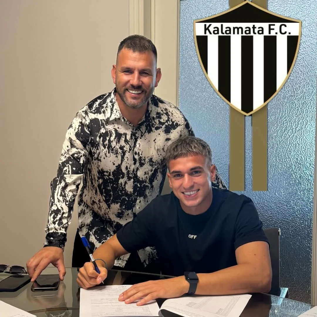 A man sitting at a desk signing papers while another man stands behind him, both smiling. There is a Kalamat F.C. crest on the textured wall behind them.