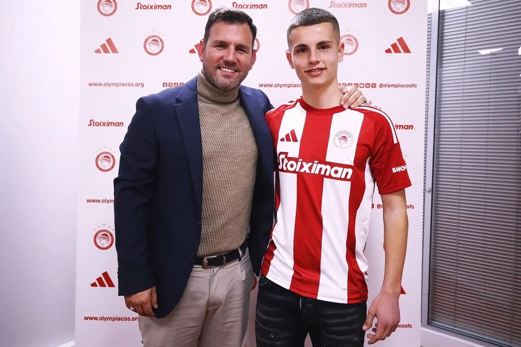 Man in a navy blazer and tie posing with a young man in a red and white Olympiacos soccer jersey at an event, in front of a backdrop with logos and social media links.