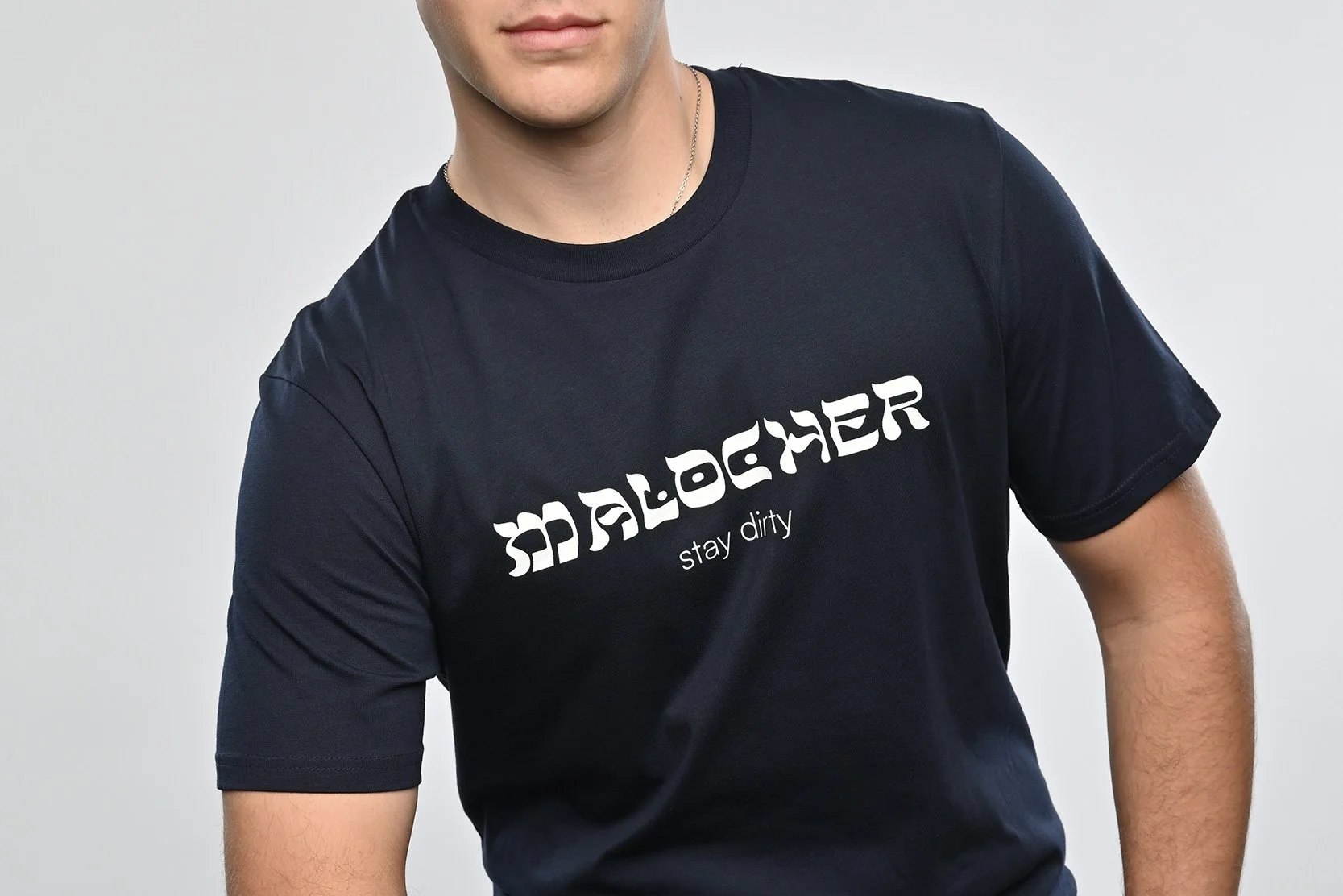 Ein junger Mann trägt ein navy blaues T-Shirt der Marke SHALOM.BERLIN mit weissem Aufdruck "MALOCHER stay dirty"