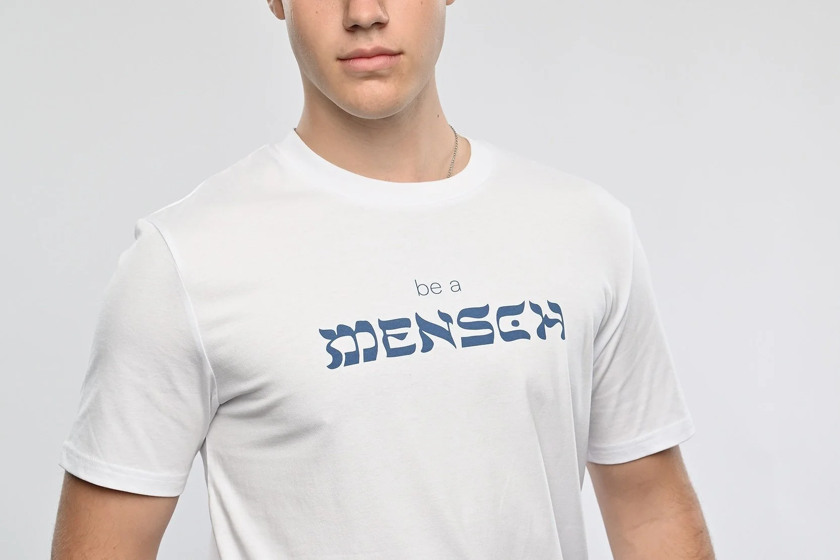 Ein junger Mann trägt ein weisses T-Shirt der Marke SHALOM.BERLIN mit blauem Aufdruck "be a MENSCH"