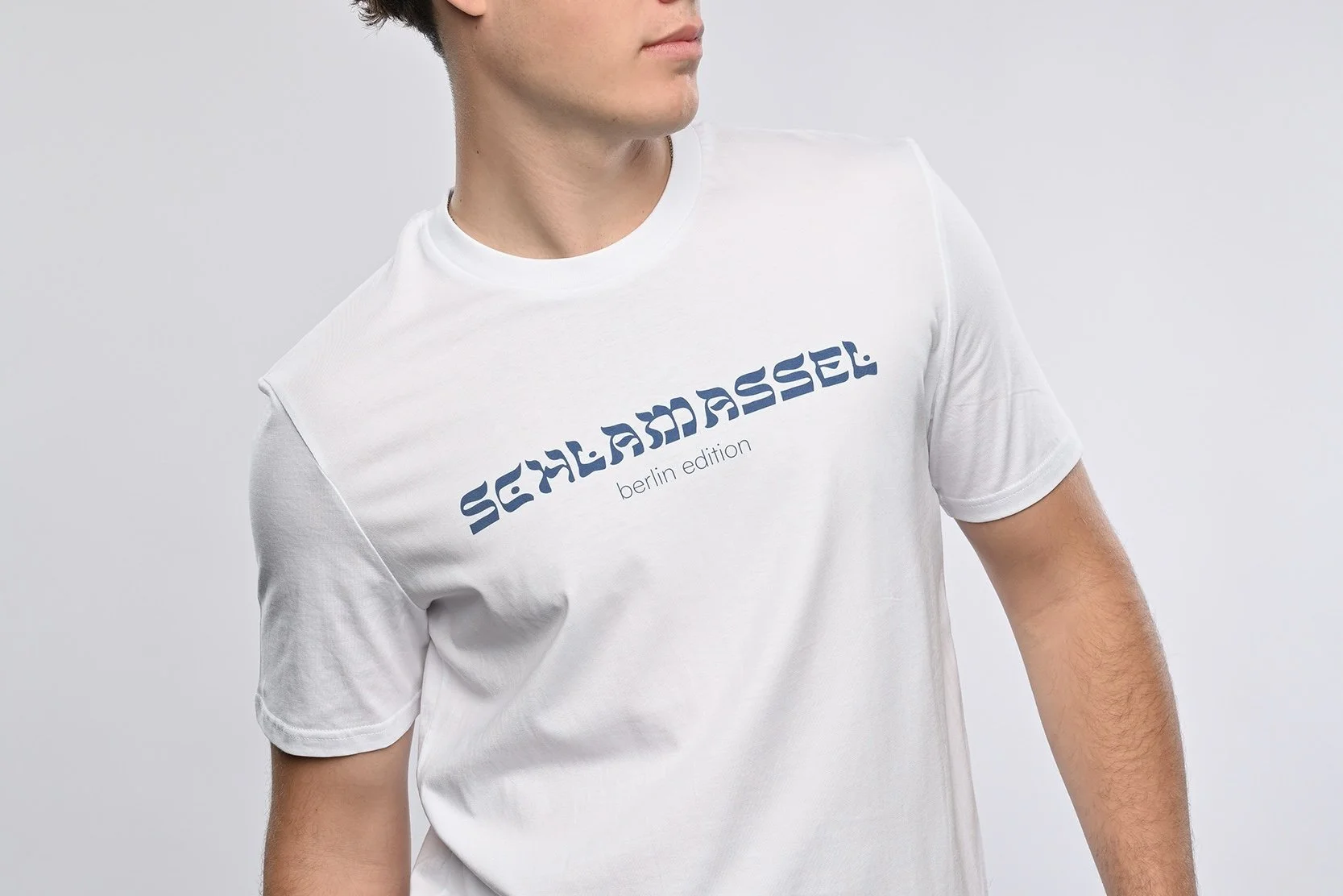 Ein junger Mann trägt ein weisses T-Shirt der Marke SHALOM.BERLIN mit blauem Aufdruck "SCHLAMASSEL berlin edition"