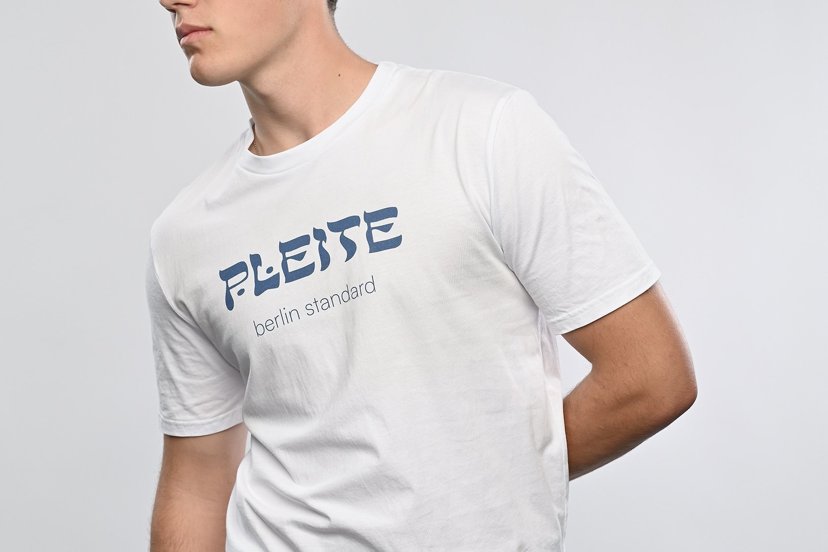 Ein junger Mann trägt ein weisses T-Shirt der Marke SHALOM.BERLIN mit blauem Aufdruck "PLEITE berlin standard"