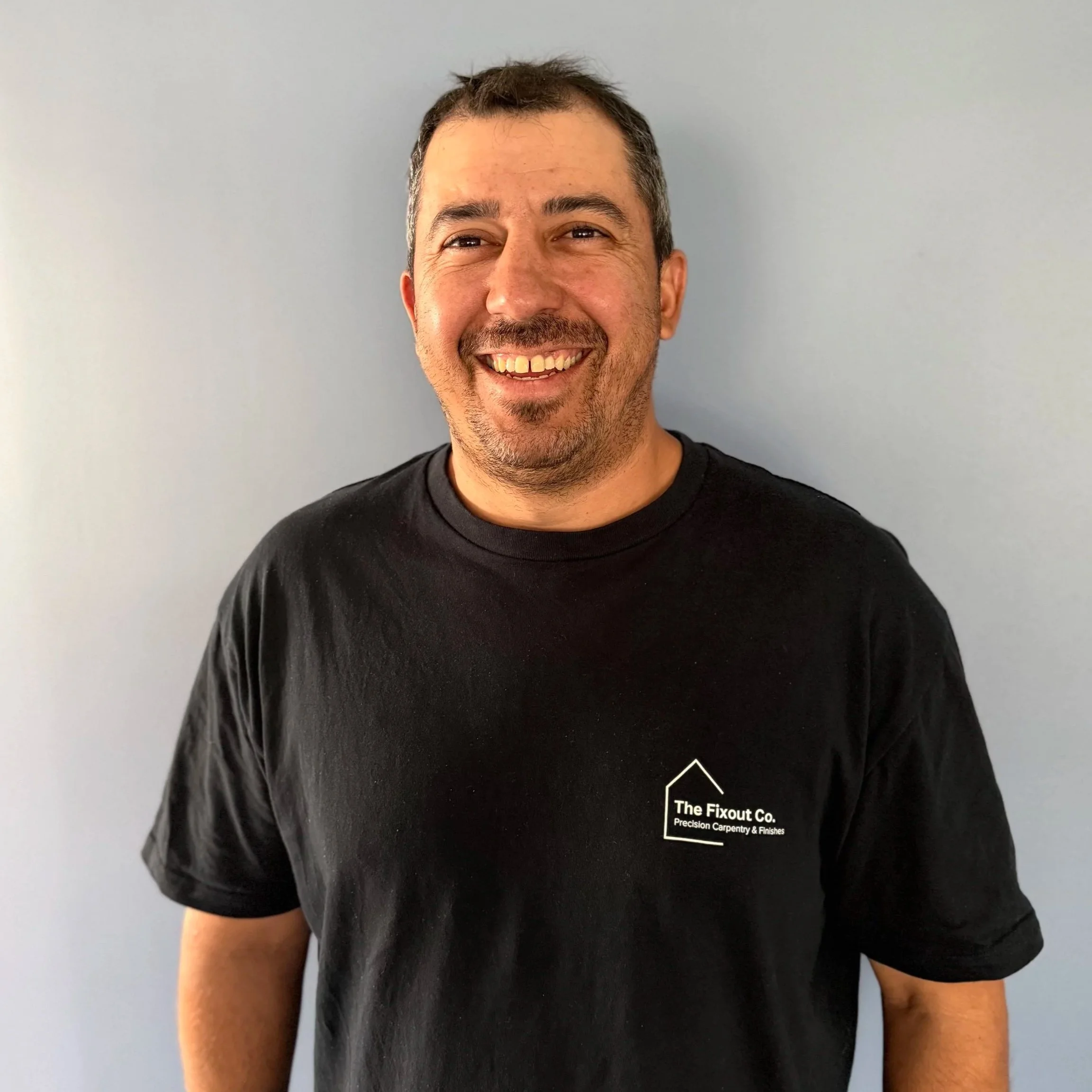 Two men smiling at a sporting event, one in a white shirt and the other in a tan jacket and plastic rain poncho, seated in a stadium with other spectators in the background.