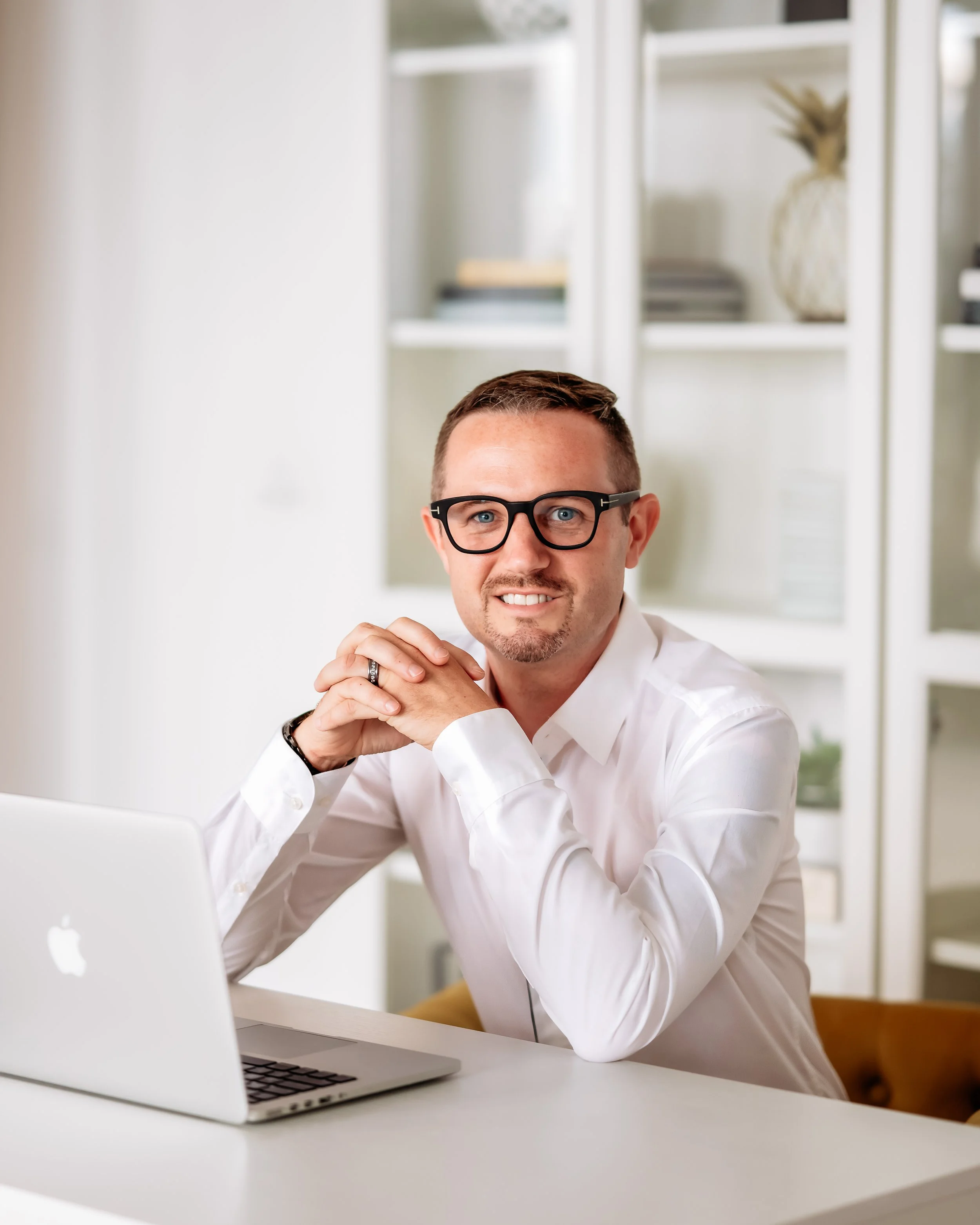 A man with glasses and a wedding ring sitting at a white desk with a silver MacBook, smiling at the camera.