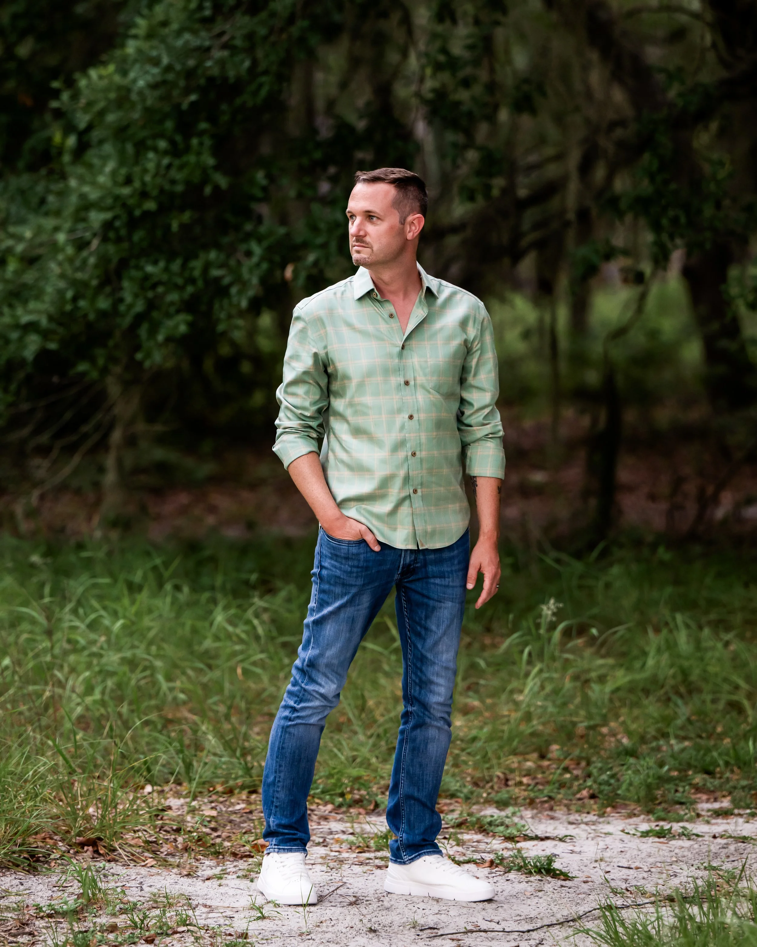 A man standing outdoors in a grassy area with trees in the background, wearing a light checkered shirt, jeans, and white sneakers, looking to his left.