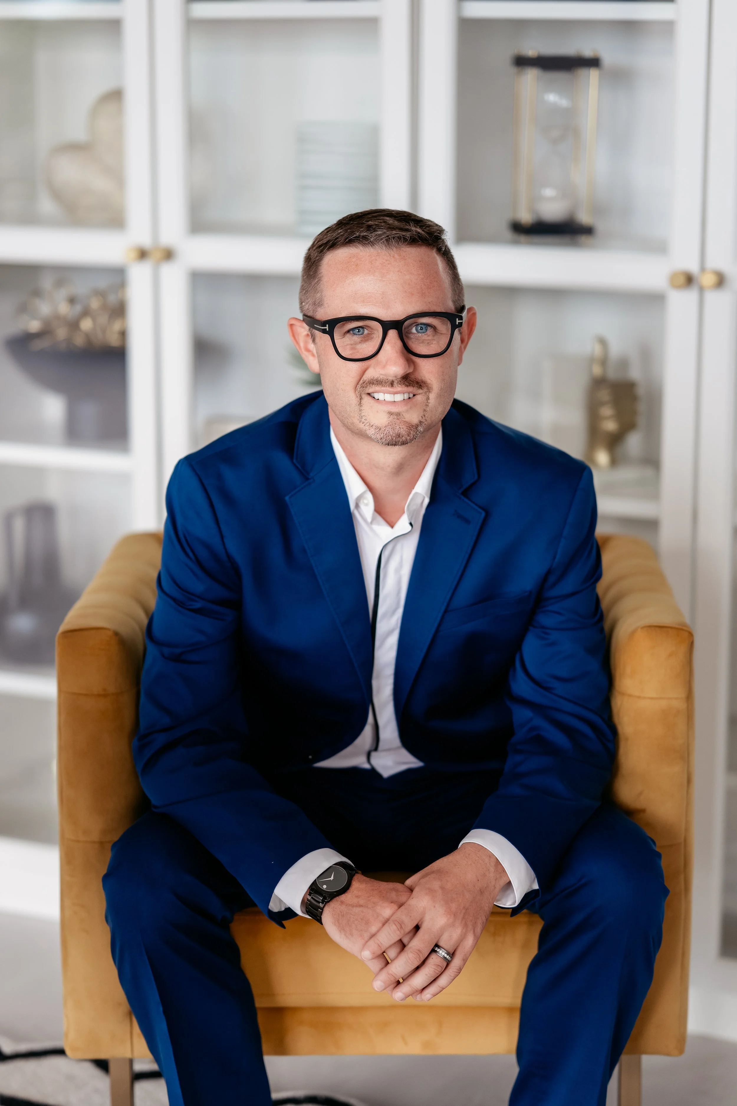A man in a blue suit with glasses, sitting on a mustard-colored chair, in front of a white cabinet with decor. He is smiling and has a black watch on his left wrist.