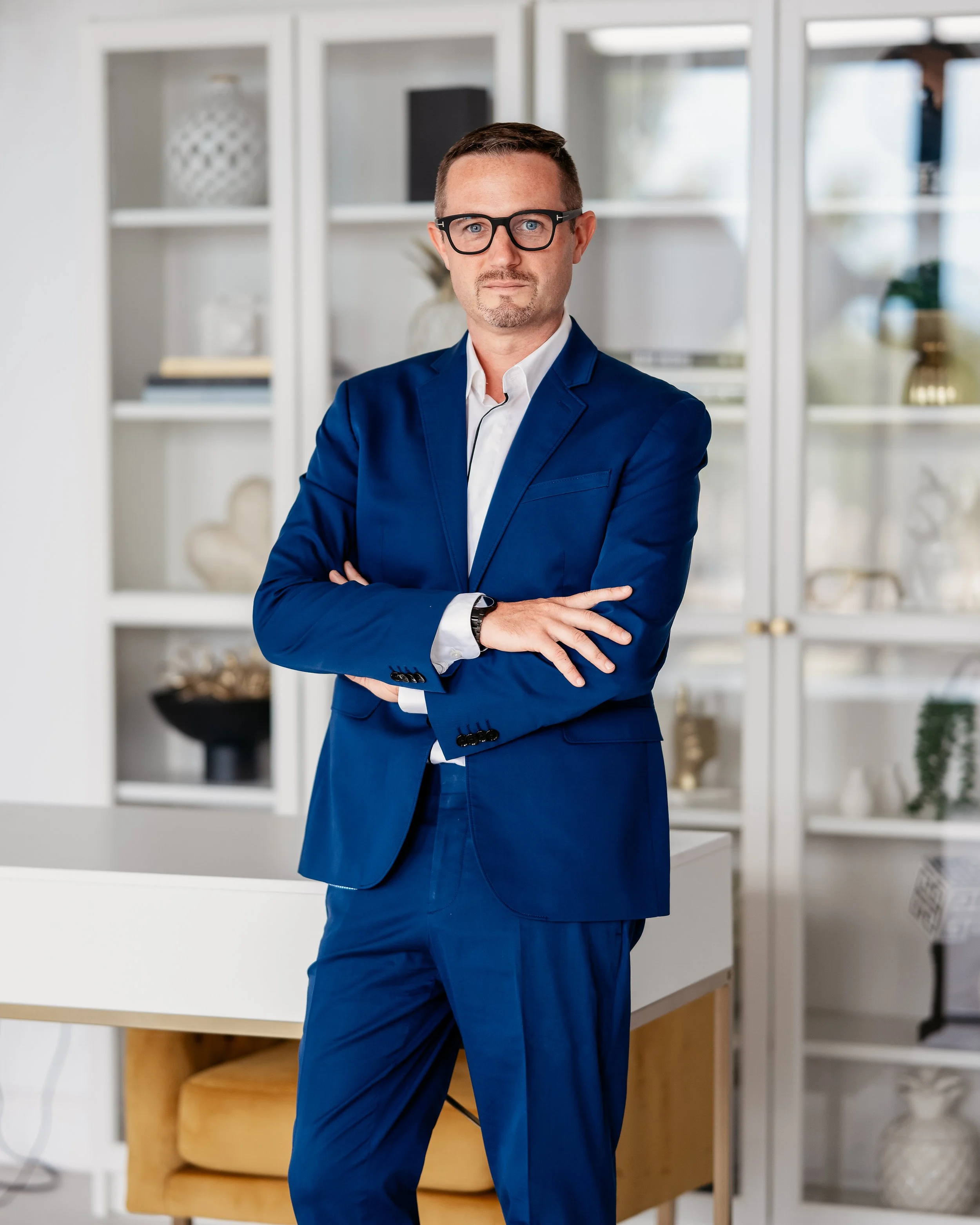 A man in a blue suit with glasses, standing with arms crossed in a modern, well-decorated office or living room.