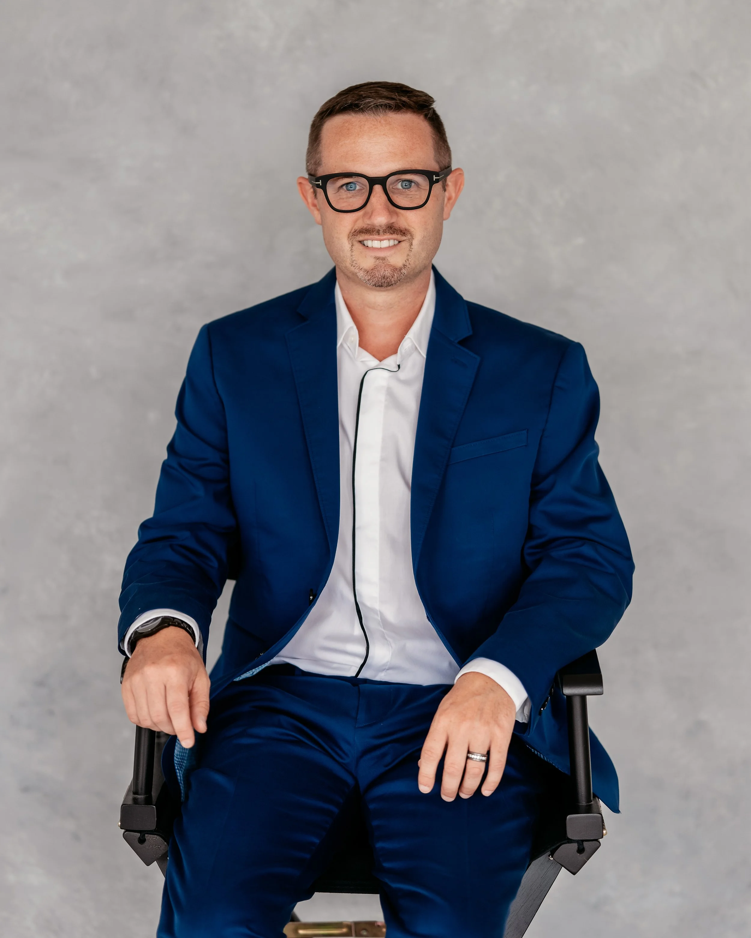 A man in a blue suit and glasses sitting on a black chair, smiling, with a neutral gray background.