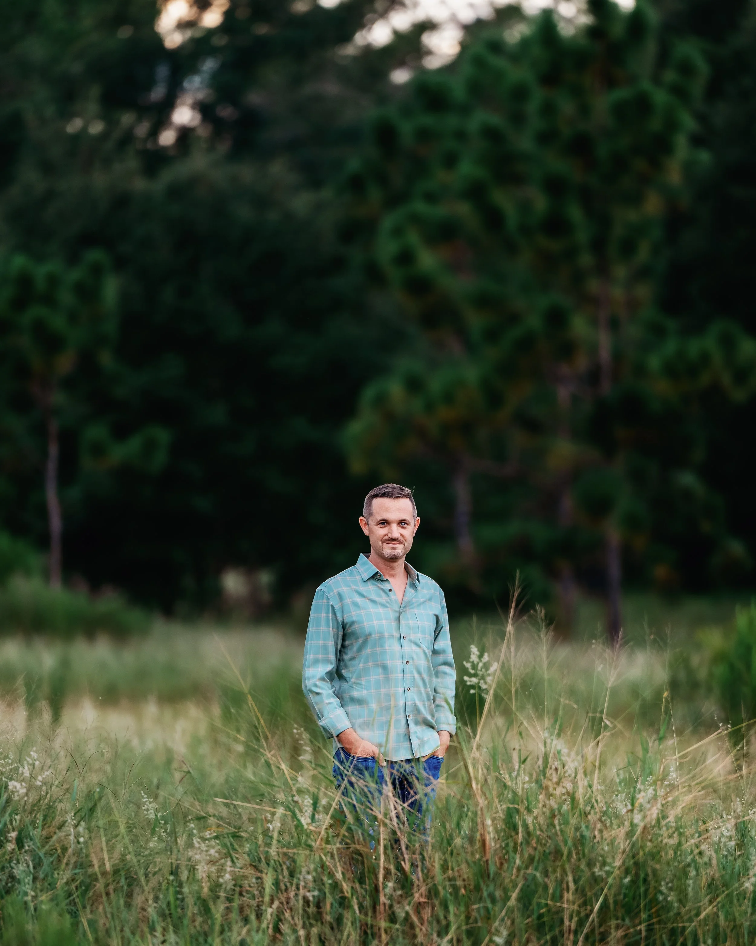 Man in checkered shirt standing in grassy field with trees in background.