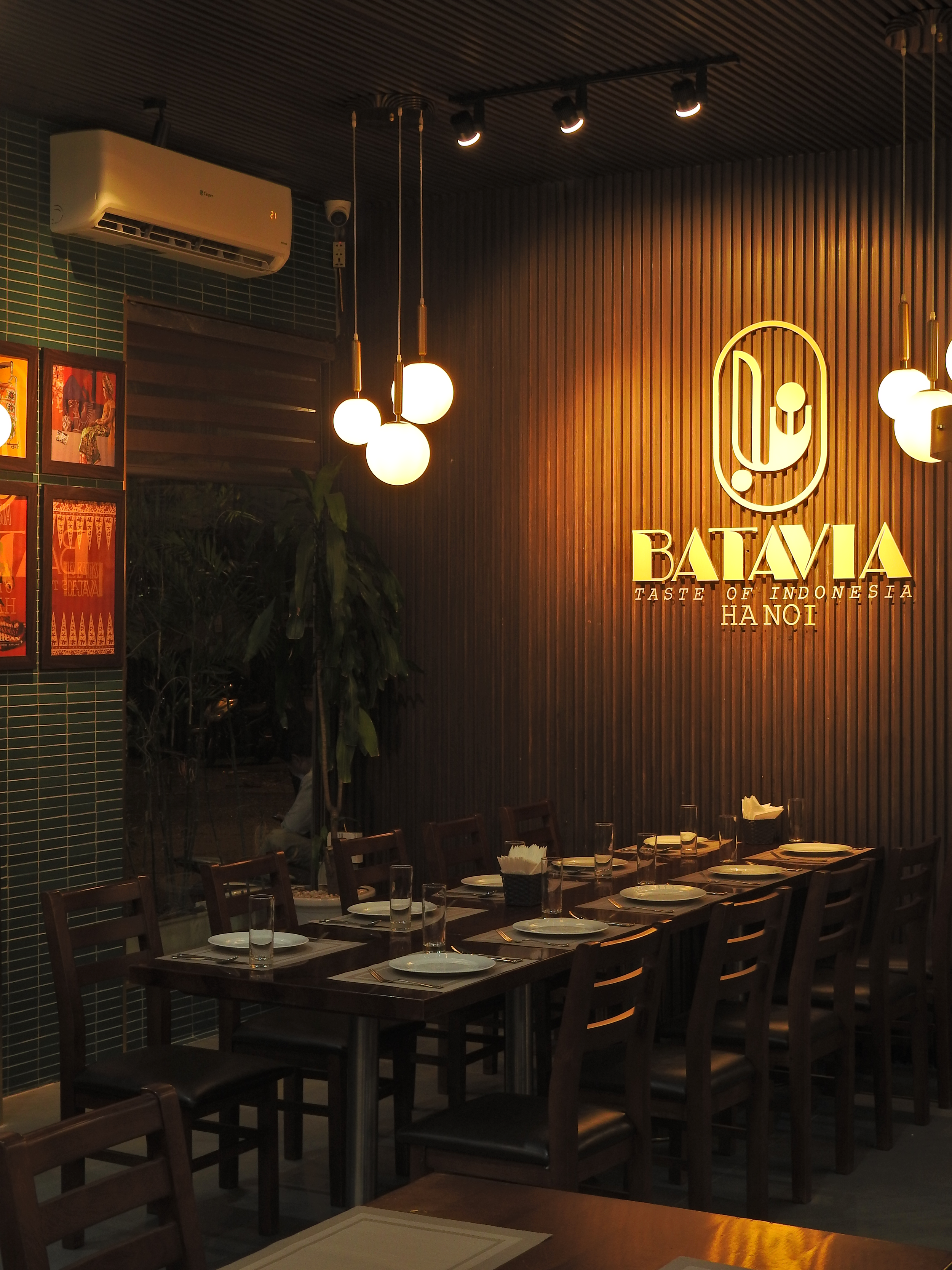 Interior of a restaurant with a long wooden table set with plates, glasses, and napkins. Decorative lights hang from the ceiling. A sign on the wooden wall reads "BATAVIA TASTE OF INDONESIA HANOI". There is a green tiled wall and some indoor plants.