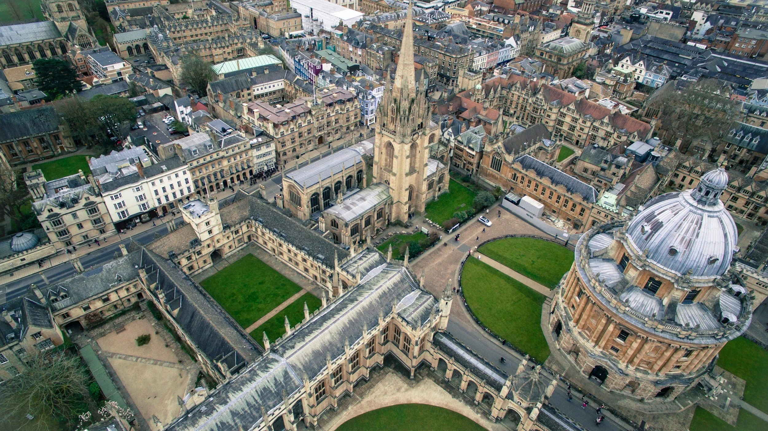 Bird's eye view of a historic European city with a large church featuring a tall spire, surrounded by medieval and modern buildings, green lawns, and narrow streets.