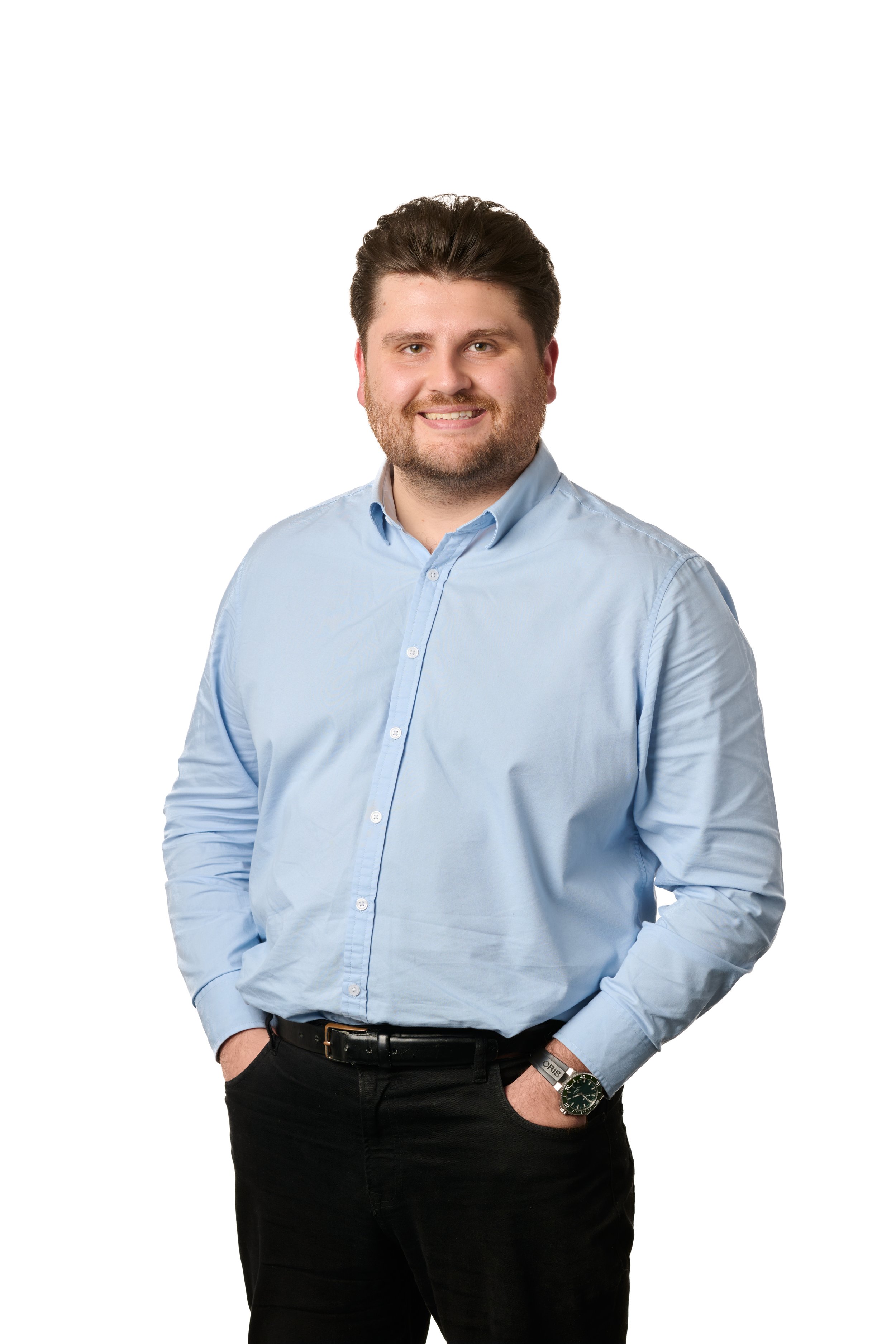 A man with dark hair, beard, and mustache wearing a light blue button-up shirt and black pants, standing against a white background, smiling with his hands in his pockets.
