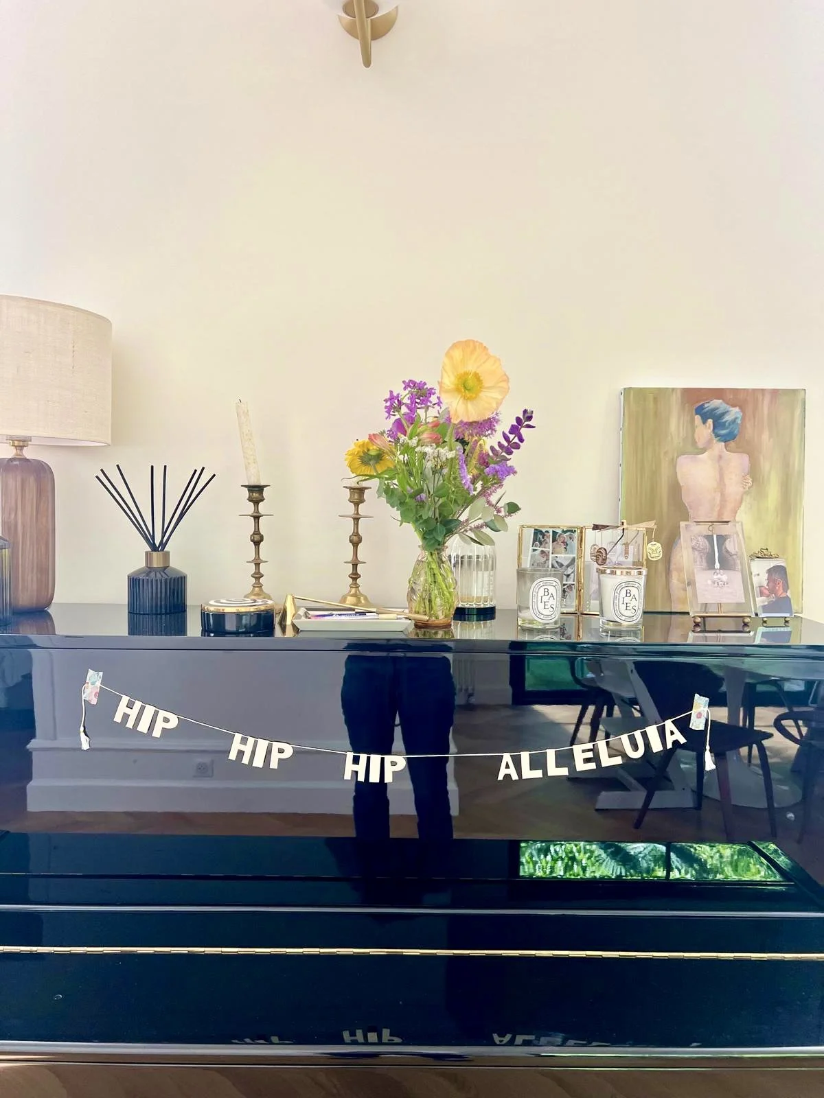 Une décoration intérieure avec un piano noir, un bouquet de fleurs colorées, des bougies, une lampe, des cadres photo, une peinture et une bannière avec la phrase "HIP HIP ALLELUJIA".