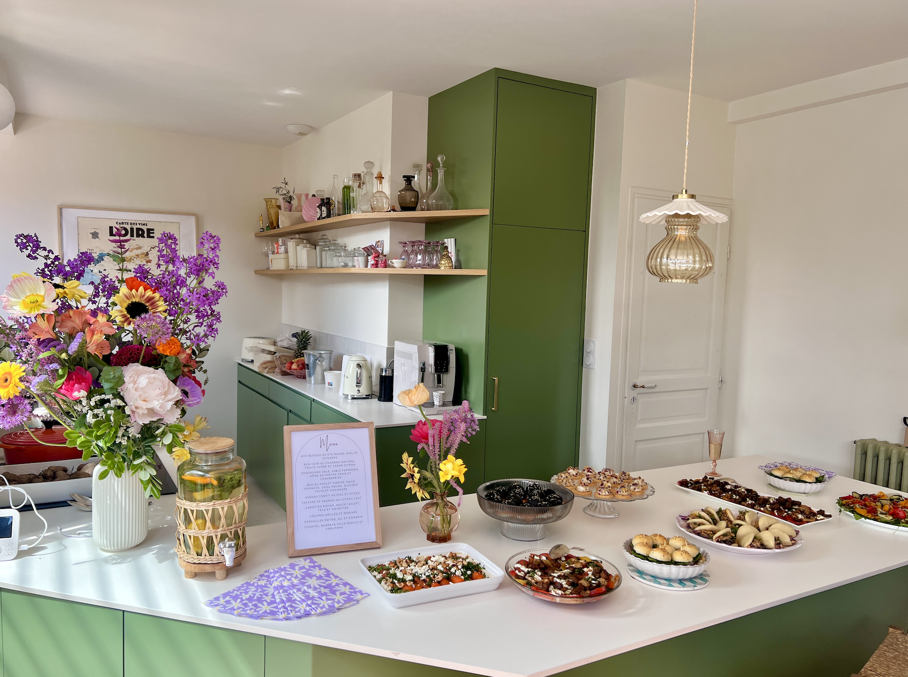 Introduction à un buffet avec un grand bouquet de fleurs colorées, une table de présentation avec des plats variés, un menu écrit dans un cadre en bois, et un décor de cuisine avec armoires vertes et étagères ouvertes.