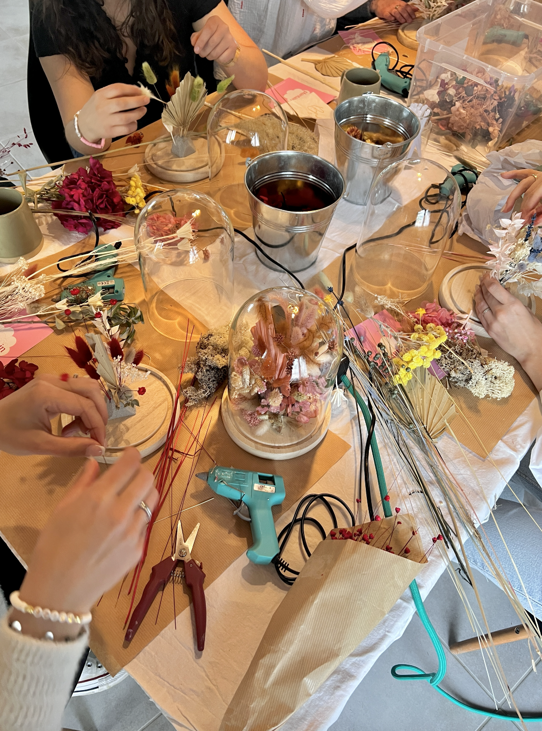 Groupe de personnes créant des arrangements floraux avec des fleurs séchées, des outils, des rubans et des décorations sur une table.