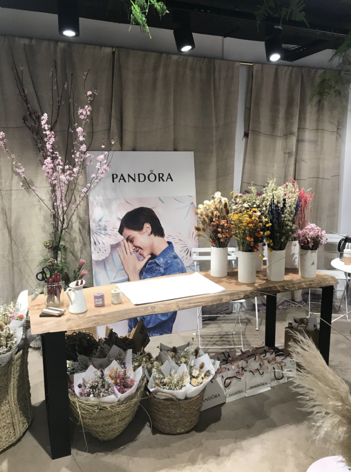 Table avec plusieurs bouquets de fleurs dans des vases blancs, une grande affiche Pandora avec une femme souriante, et quelques objets déco et cadeaux, dans un espace intérieur.