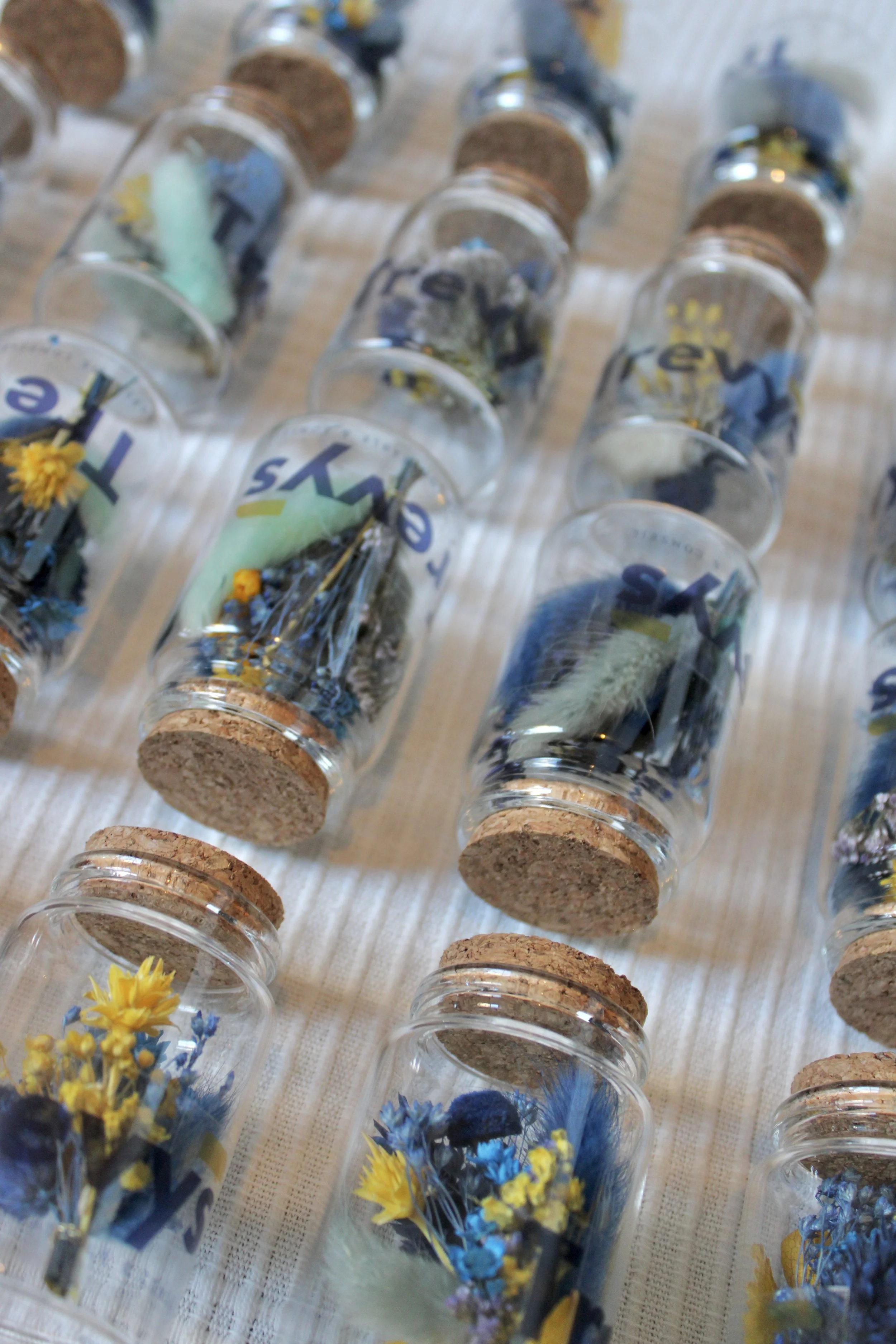 Vases en verre avec des fleurs séchées jaunes, bleues et blanches, certains contenant des petits objets, alignés sur une surface en tissu.