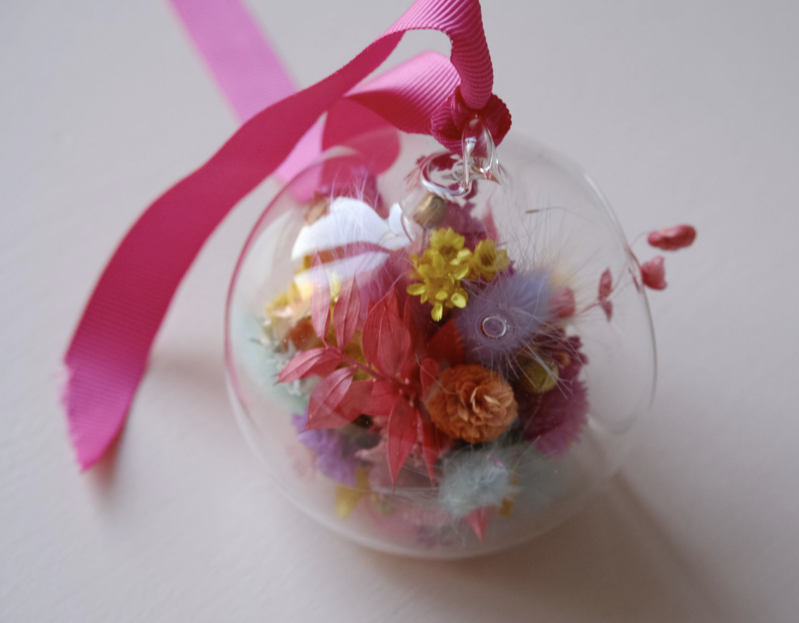 Ornement en forme de boule transparente contenant des fleurs colorées avec un ruban rose attaché au sommet.